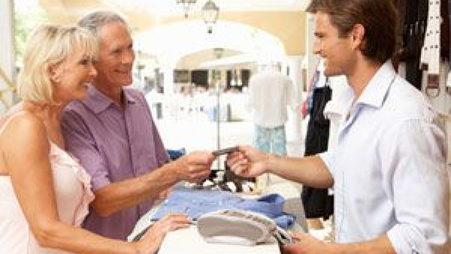 An older couple stands at a store counter, smiling as the man hands a credit card to a young male cashier. Clothes and a credit card terminal are visible on the counter in a bright, open shop.
