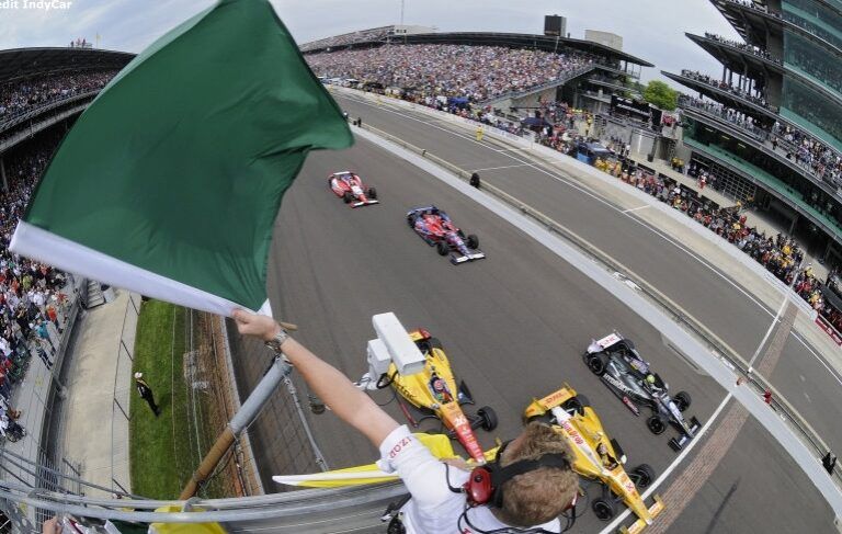 A race official waves a green flag to start an IndyCar race, with cars speeding down the track and a packed grandstand of spectators in the background.