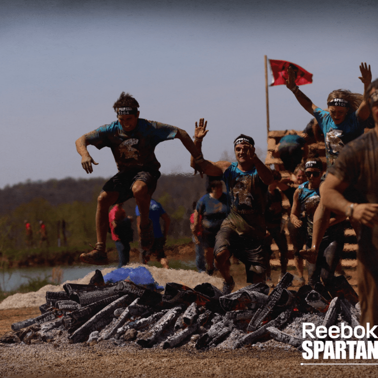 Group of people in athletic gear and headbands leap over burning logs during a muddy outdoor obstacle course race, with a Reebok Spartan Race logo in the bottom right corner.