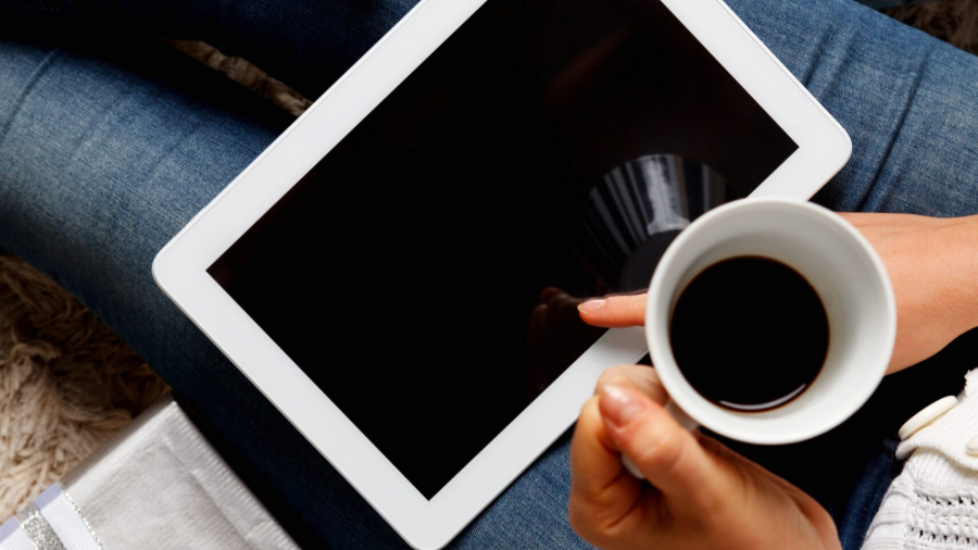 Person sitting with wrapped gifts around, holding a cup of coffee and a tablet on their lap, with a credit card visible, suggesting online shopping or gift planning.