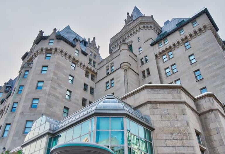 A low-angle view of a large historic stone building with castle-like architecture, featuring turrets, peaked roofs, and numerous windows. Modern glass windows are visible at the ground level.
