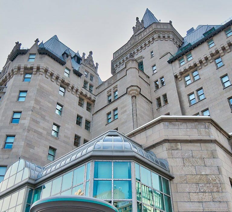 Stone building with castle-like architecture, featuring turrets, pointed roofs, and decorative details, set against a pale sky. Lower part has modern glass windows and a metallic roof accent.