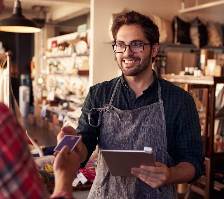 A smiling shop worker in an apron holds a tablet while taking a payment from a customer with a credit card inside a cozy store with shelves of home goods.
