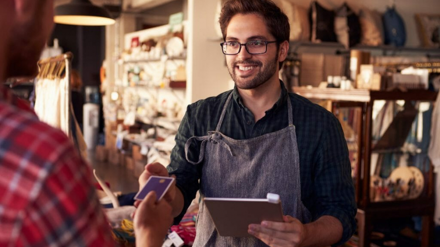 A smiling shop worker in an apron holds a tablet while taking a payment from a customer with a credit card inside a cozy store with shelves of home goods.