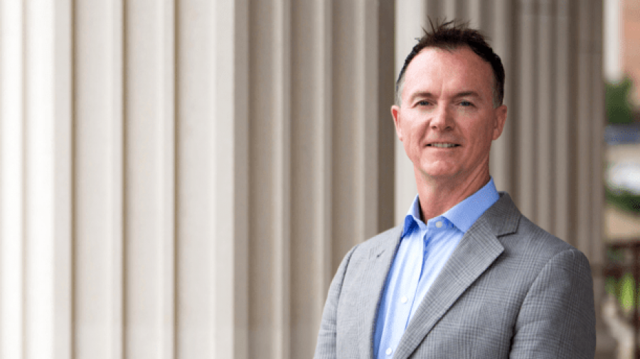 A man in a light gray suit and blue shirt stands outside in front of tall white columns, smiling at the camera.