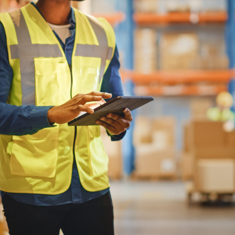A person wearing a yellow safety vest uses a tablet in a warehouse with shelves and cardboard boxes in the background.