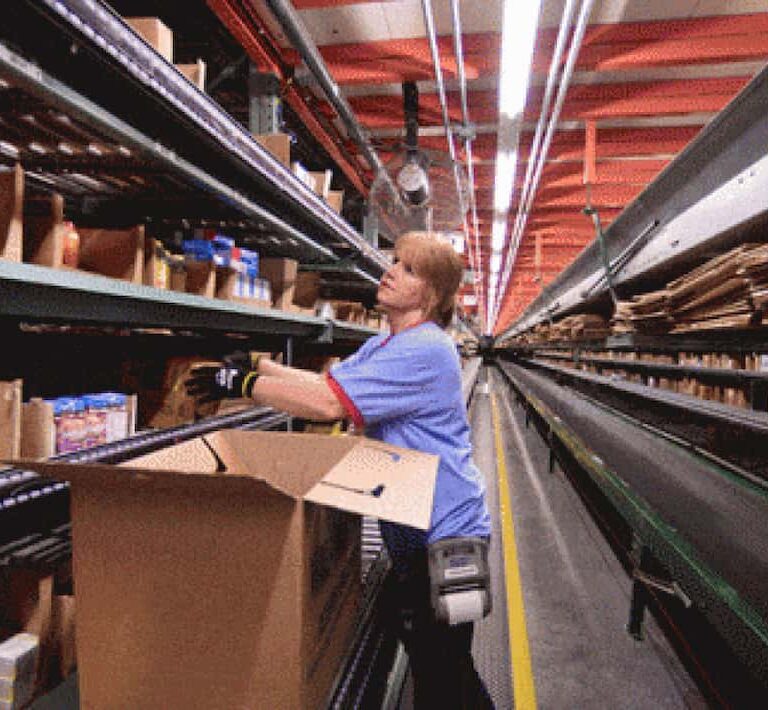 A worker in a warehouse aisle selects items from shelves and places them into a large cardboard box on a conveyor belt, preparing orders for shipment.