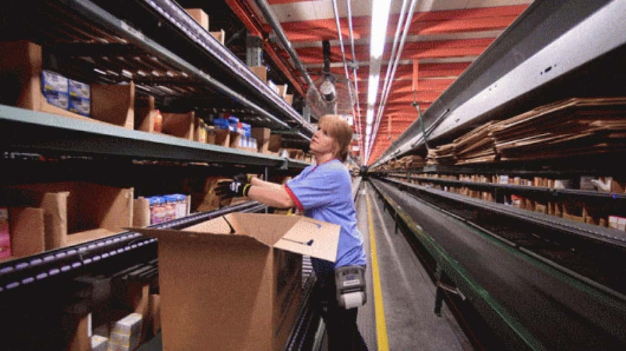 A worker in a warehouse aisle selects items from shelves and places them into a large cardboard box on a conveyor belt, preparing orders for shipment.