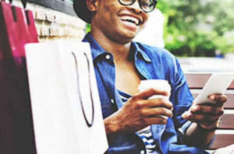 A person sitting on a bench, smiling while holding a coffee cup and using a tablet, with shopping bags beside them. They are wearing glasses, a hat, and a denim shirt.