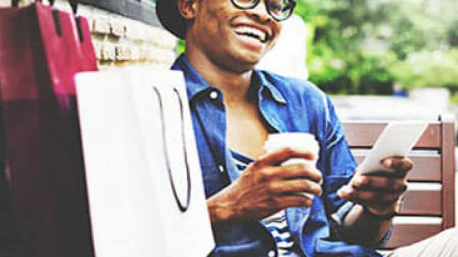 A person sitting on a bench, smiling while holding a coffee cup and using a tablet, with shopping bags beside them. They are wearing glasses, a hat, and a denim shirt.