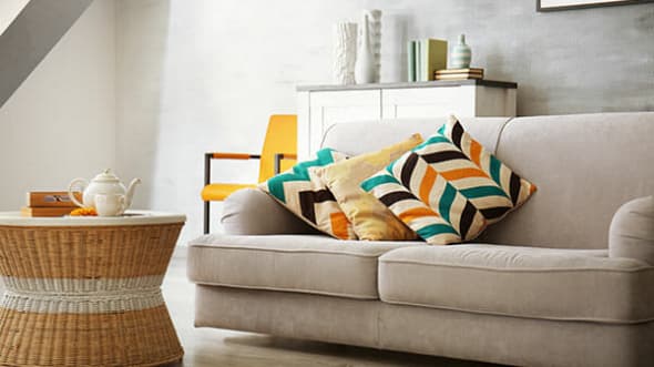 A beige sofa with colorful chevron and yellow pillows sits next to a wicker coffee table with a white teapot and cup. In the background, there’s a white cabinet, an orange chair, and decorative vases.