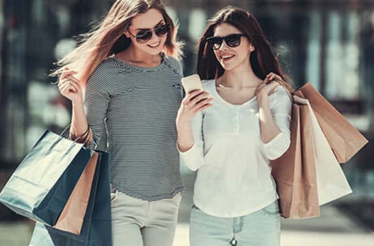Two young women wearing sunglasses smile while walking outdoors, holding shopping bags and looking at a smartphone together.