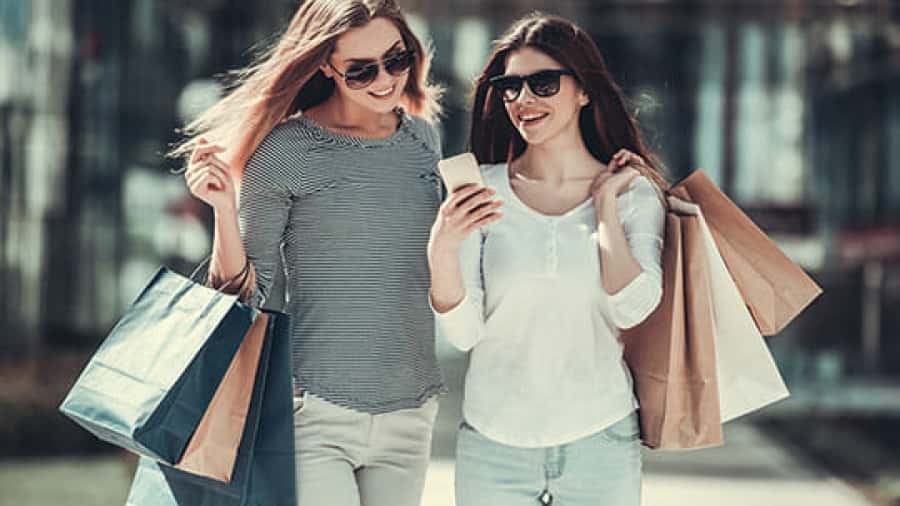 Two young women wearing sunglasses smile while walking outdoors, holding shopping bags and looking at a smartphone together.