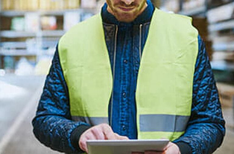 A person wearing a yellow safety vest uses a tablet in a warehouse setting, surrounded by shelves and boxes.