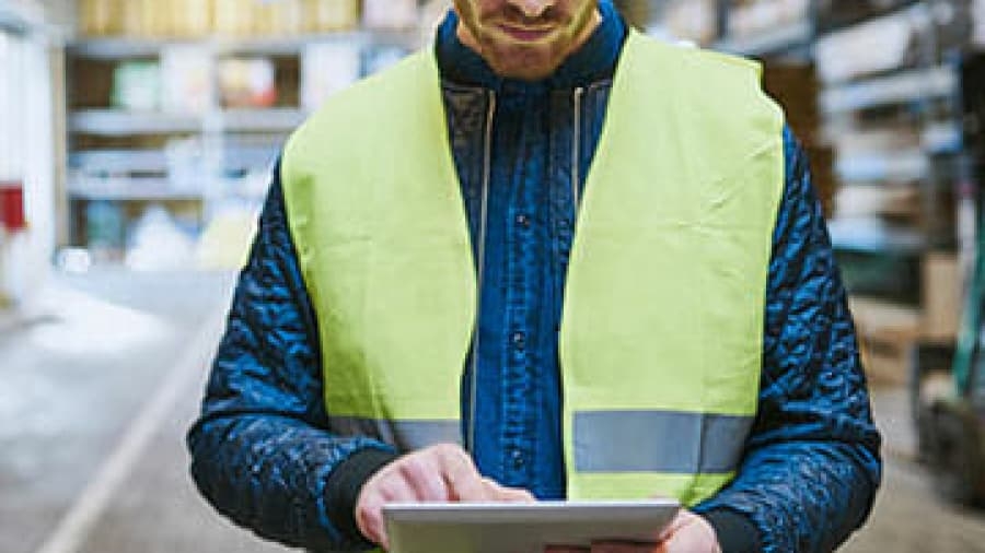 A person wearing a yellow safety vest uses a tablet in a warehouse setting, surrounded by shelves and boxes.