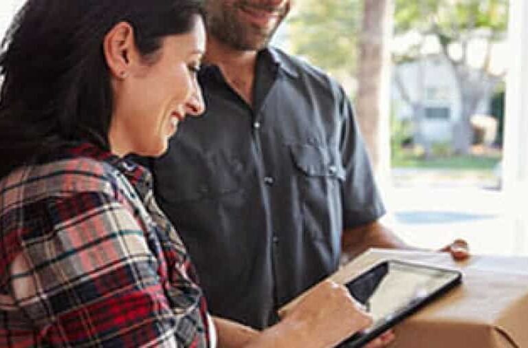 A woman smiles as she signs for a package on a tablet held by a delivery person at her door, both standing indoors with sunlight coming through a window behind them.