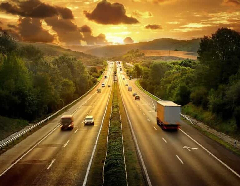 A highway stretches into the distance, surrounded by green trees and hills, with cars and a truck driving under a dramatic golden sunset sky filled with clouds.