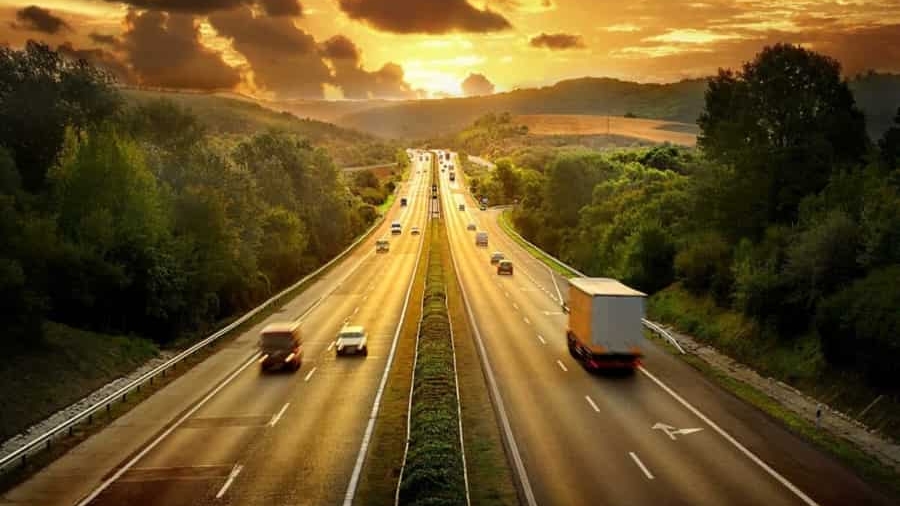 A highway stretches into the distance, surrounded by green trees and hills, with cars and a truck driving under a dramatic golden sunset sky filled with clouds.