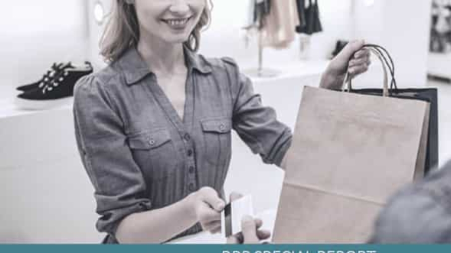 A smiling woman at a store counter hands a paper shopping bag to a customer while the customer offers a credit card for payment. Clothing and shoes are visible in the background.