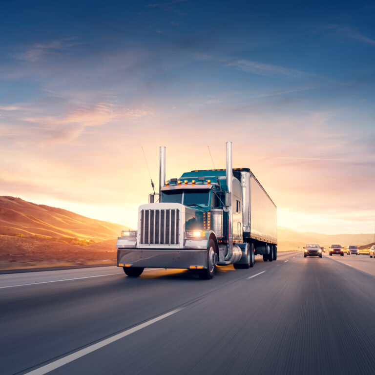A large semi-truck drives down an open highway at sunset, with hills in the background and several cars traveling in adjacent lanes under a colorful sky.