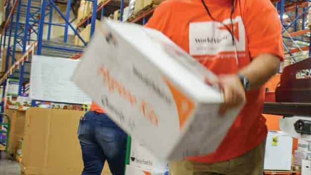 A person wearing an orange shirt holds a large white box labeled Hygiene Kit in a warehouse filled with shelves and packed boxes. Another person in jeans and an orange shirt works in the background.