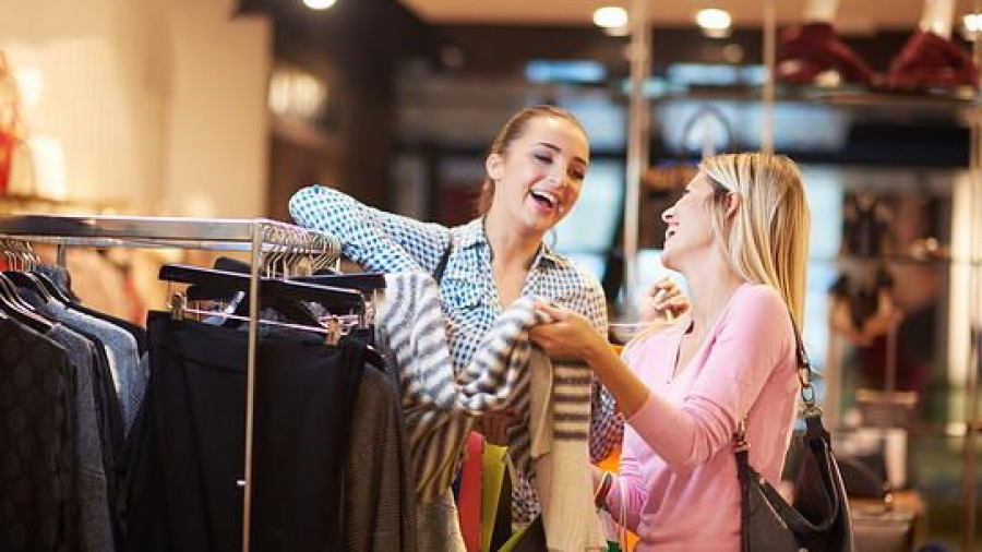 Two women smiling and laughing while shopping for clothes in a brightly lit store, holding and looking at a striped sweater near a rack of clothing.