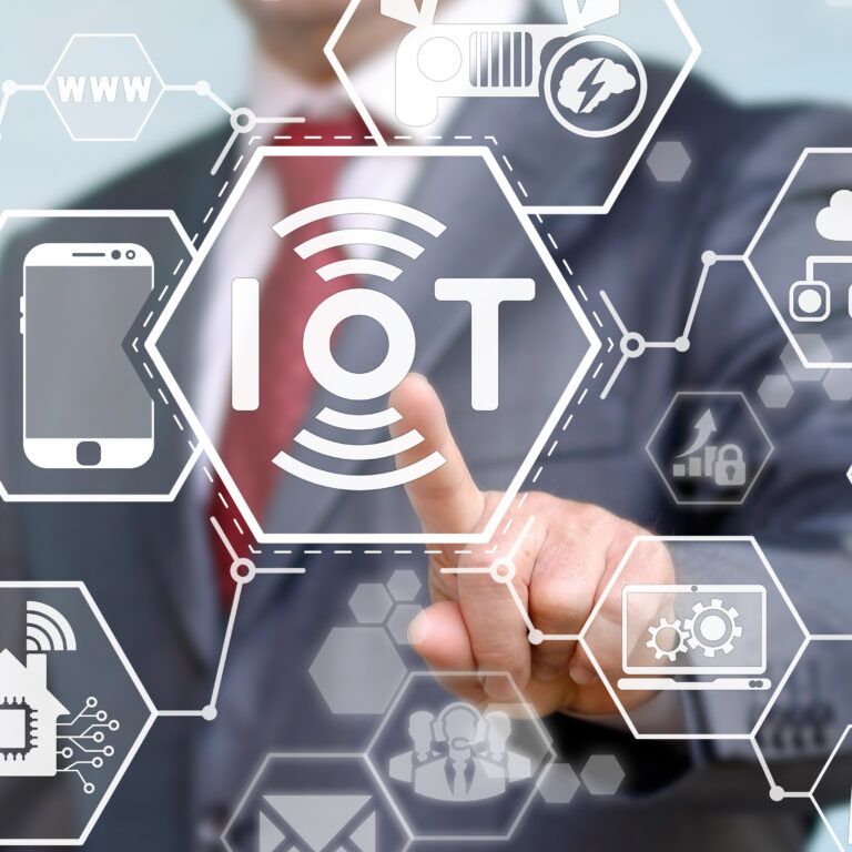 A person in a suit touches a transparent screen with “IoT” at the center, surrounded by icons representing smart devices and technologies, symbolizing the Internet of Things concept.