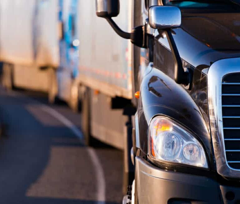 Close-up view of the front of a black semi-truck driving on a curved road, with other trucks visible in the background, all traveling in the same direction.