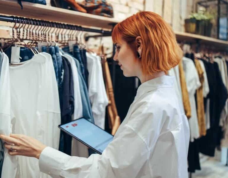A woman with short red hair in a white shirt holds a tablet while inspecting clothes on a rack in a boutique clothing store.