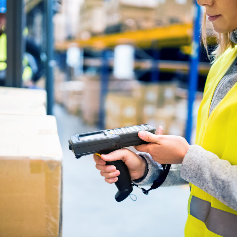 A woman wearing a yellow safety vest uses a handheld barcode scanner to scan boxes in a warehouse. Shelves with more boxes are visible in the background.