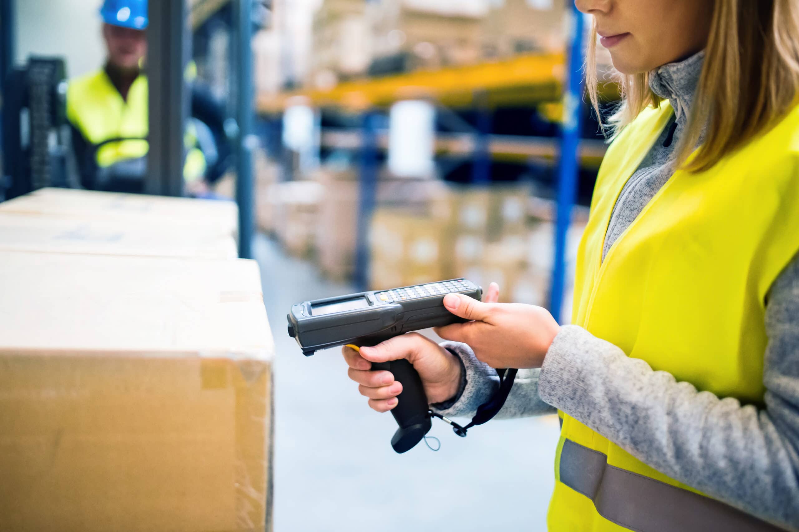 A woman wearing a yellow safety vest uses a handheld barcode scanner to scan boxes in a warehouse. Shelves with more boxes are visible in the background.