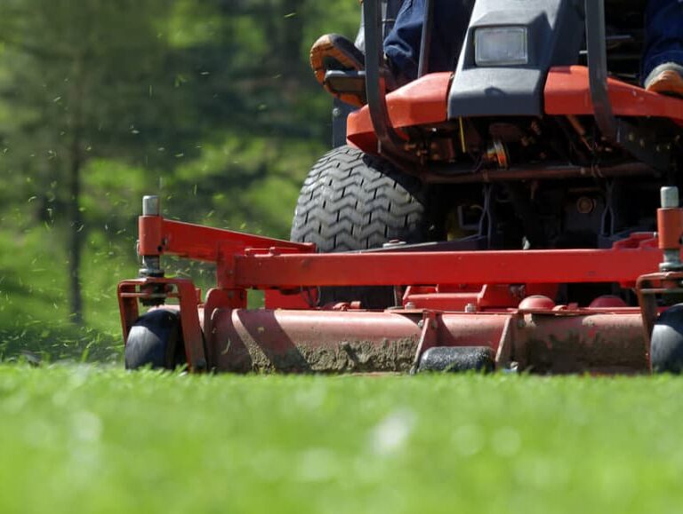 Close-up view of a person riding a red lawn mower, cutting green grass on a sunny day. Grass clippings are visible flying from the mower, with trees and greenery in the blurred background.
