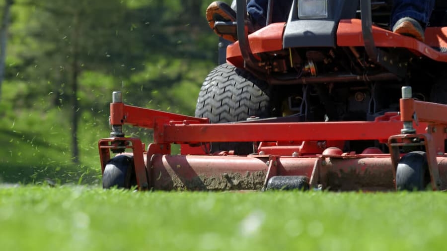 Close-up view of a person riding a red lawn mower, cutting green grass on a sunny day. Grass clippings are visible flying from the mower, with trees and greenery in the blurred background.