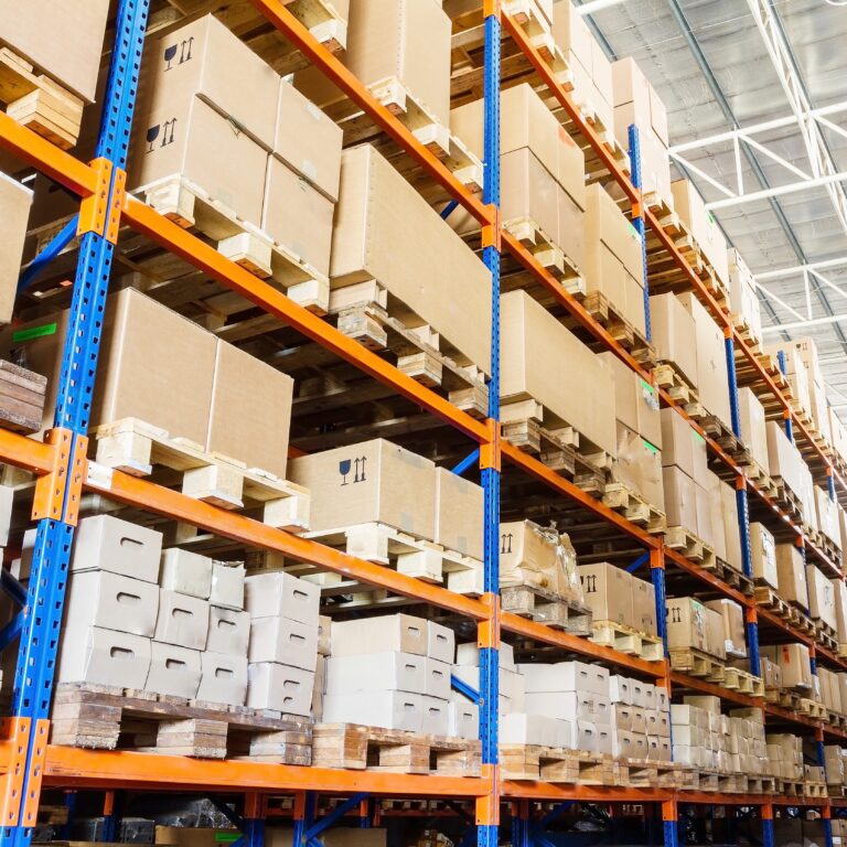 Large warehouse with tall blue and orange metal shelving filled with cardboard boxes and packages stacked on wooden pallets, under a high ceiling with natural light.