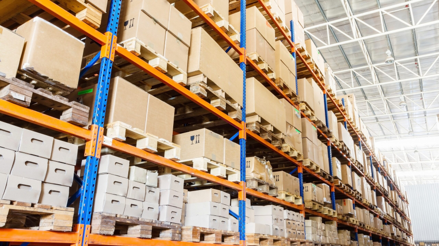 Large warehouse with tall blue and orange metal shelving filled with cardboard boxes and packages stacked on wooden pallets, under a high ceiling with natural light.