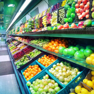 A supermarket produce section with rows of neatly organized fresh fruits and vegetables in baskets and crates, including apples, oranges, lemons, pears, and more, under bright lighting.