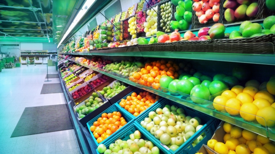 A supermarket produce section with rows of neatly organized fresh fruits and vegetables in baskets and crates, including apples, oranges, lemons, pears, and more, under bright lighting.