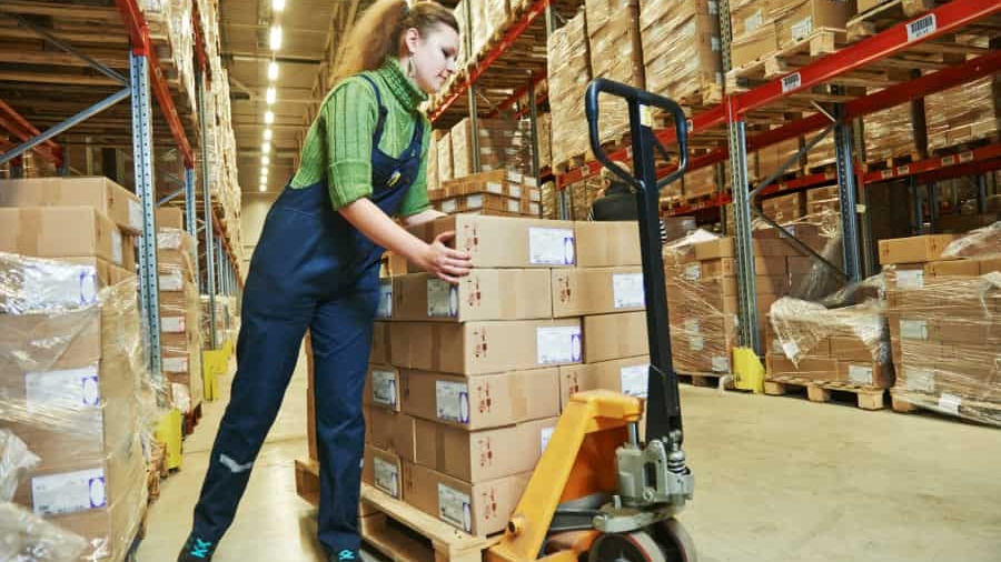 A worker in overalls moves a pallet loaded with cardboard boxes using a manual pallet jack inside a large warehouse filled with shelves of stacked boxes.