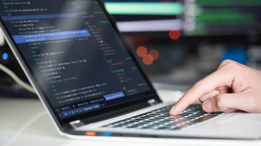 A close-up of a hand typing on a laptop keyboard, with programming code displayed on the screen. In the blurred background, additional monitors show more lines of code.