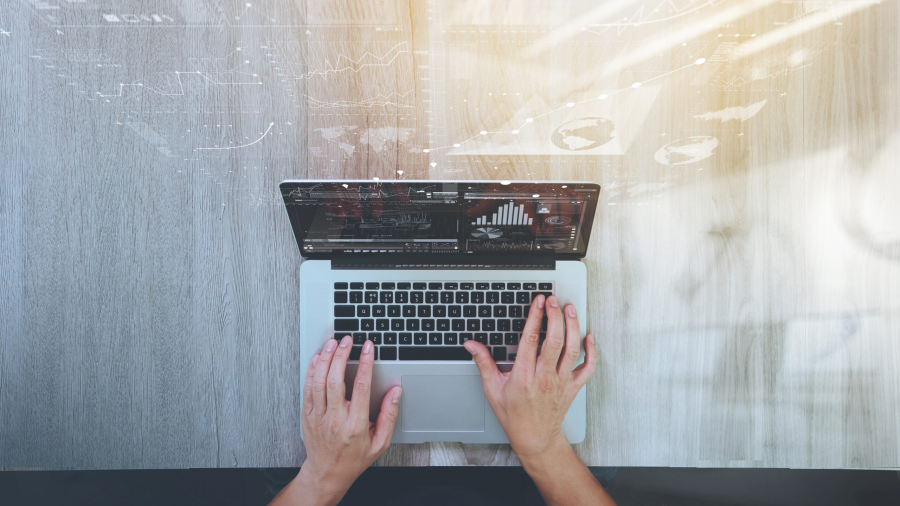 A person types on a laptop at a wooden desk, with data charts and graphs digitally overlaid above the screen, suggesting analysis or business activity. Sunlight shines on the right side of the workspace.