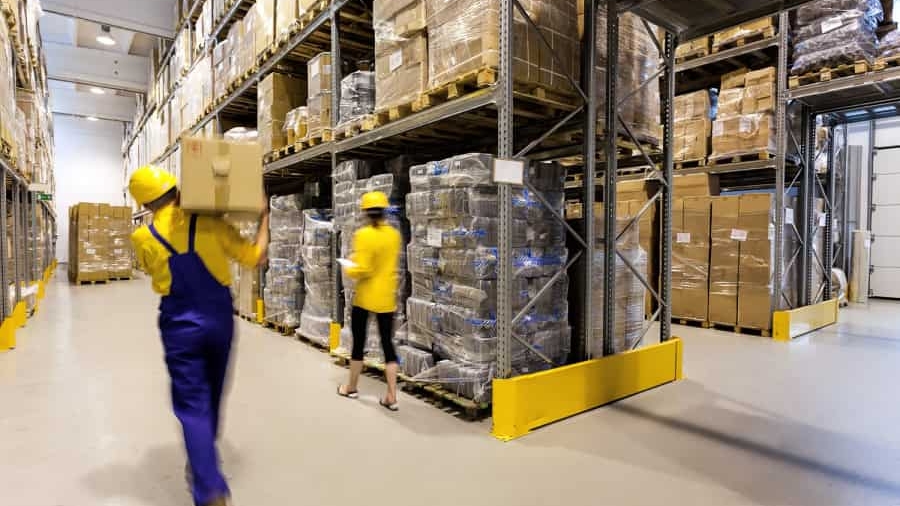 Two workers in yellow helmets and uniforms move boxes in a large warehouse with tall shelves filled with stacked cartons and pallets wrapped in plastic. The environment is brightly lit and organized.