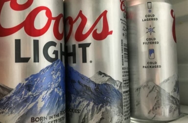 Three cans of Coors Light beer with mountain graphics are placed close together, with two in the foreground and one in the background inside a refrigerator.