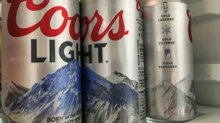 Three cans of Coors Light beer with mountain graphics are placed close together, with two in the foreground and one in the background inside a refrigerator.
