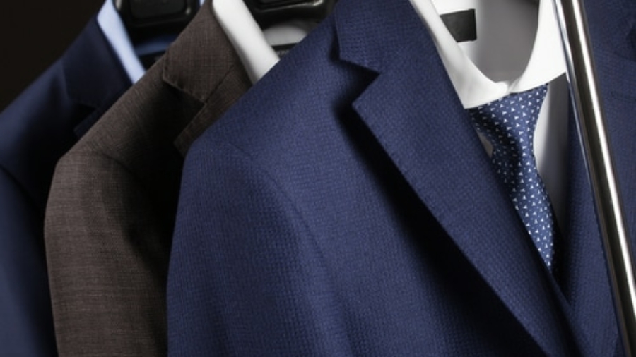 Three suits hanging on hangers, featuring a blue suit with a white shirt and dotted tie in front, followed by a brown and a dark blue suit in the background.