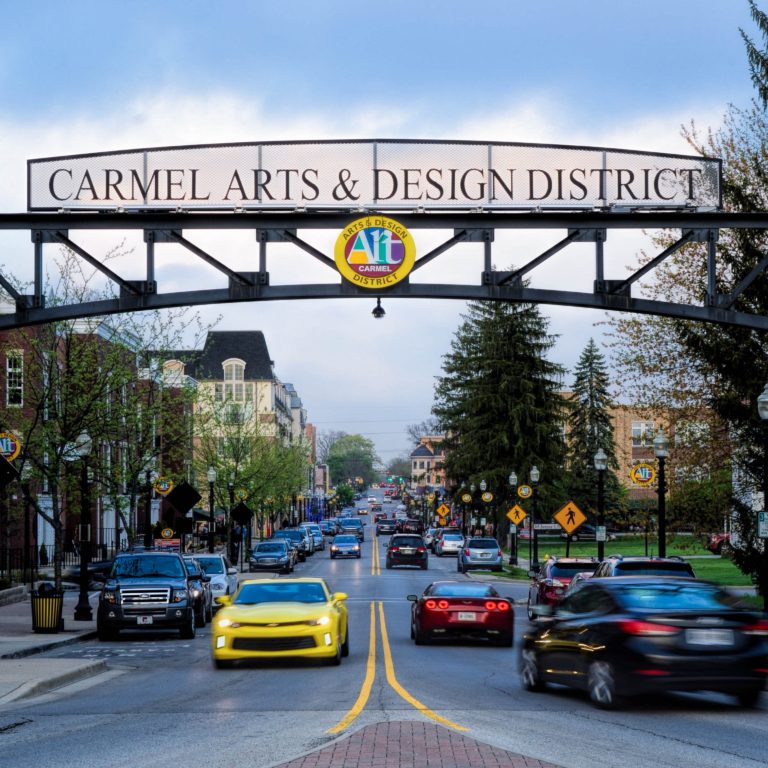 A street view of the Carmel Arts & Design District entrance with a large arched sign overhead, cars driving by, trees, brick pillars, and city buildings visible in the background.