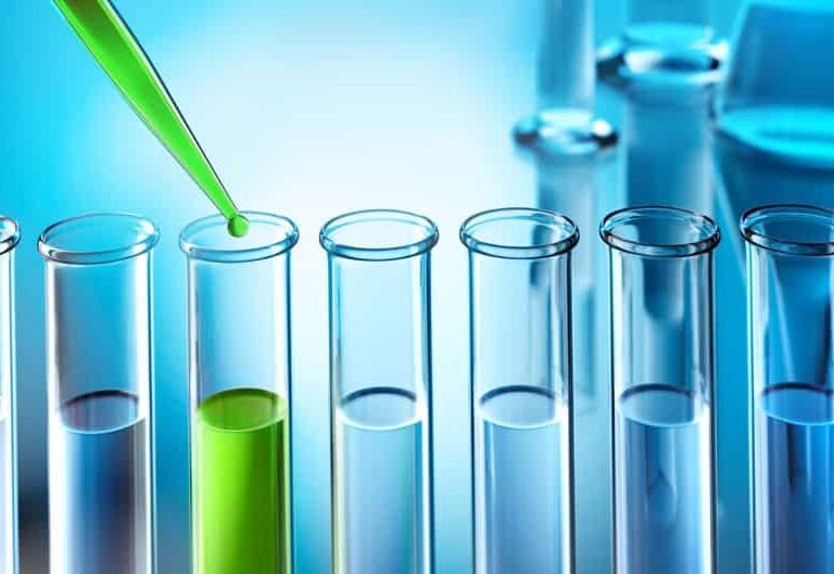 A green liquid is being dispensed from a pipette into one of several clear test tubes, with only one tube containing the green liquid. The background is blue and features blurred laboratory glassware.