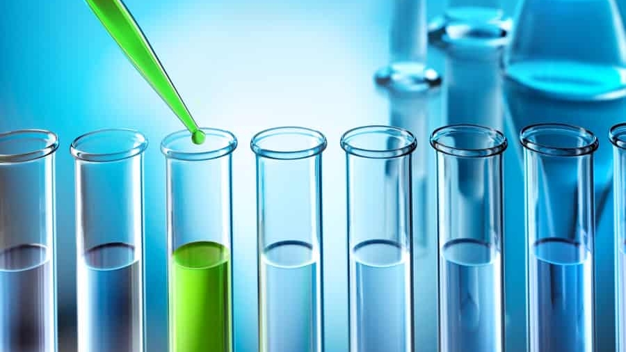 A green liquid is being dispensed from a pipette into one of several clear test tubes, with only one tube containing the green liquid. The background is blue and features blurred laboratory glassware.