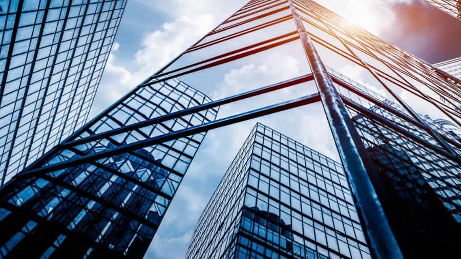 Tall glass skyscrapers reflecting the sky and clouds, viewed from below, with sunlight shining between the buildings, creating a modern and urban cityscape.