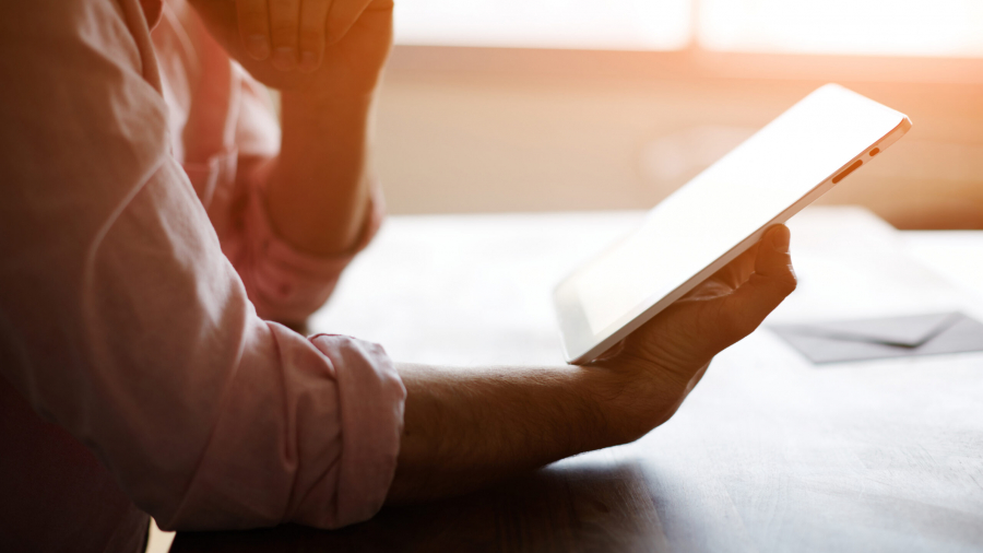 A person wearing a light pink shirt sits at a table, holding a white tablet in one hand and resting their chin on the other hand, with sunlight streaming in from a window in the background.