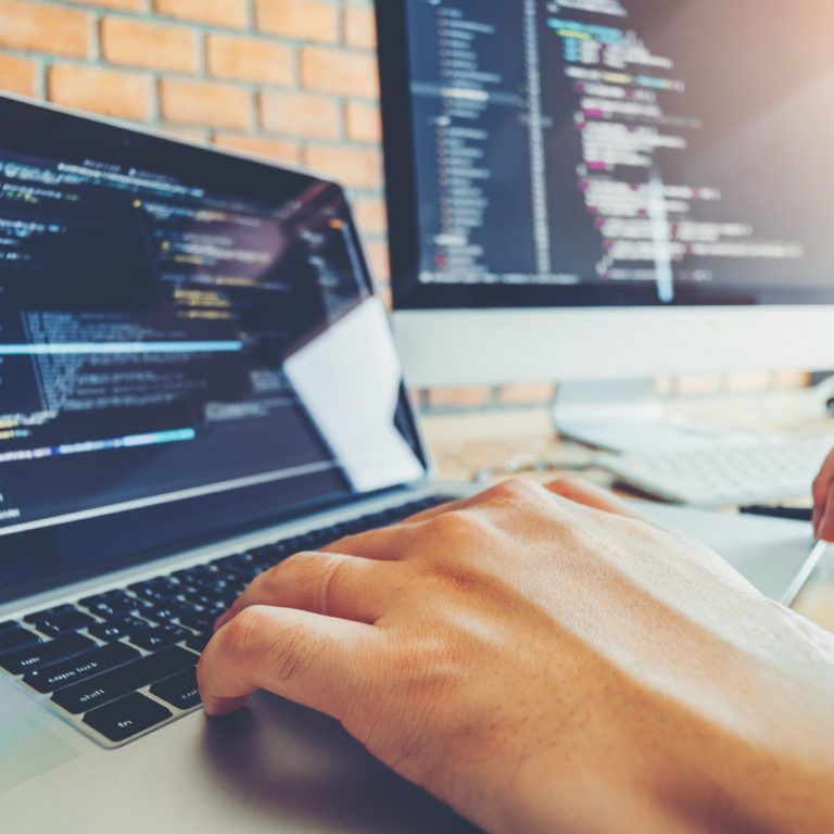 A person types on a laptop with code displayed on the screen. Two large monitors, also showing code, are on a desk in the background—a modern workspace ideal for tech roles like those at enVista Careers.